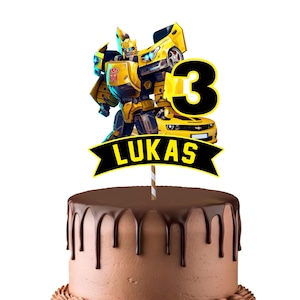 Puede incluir: Un pastel de chocolate con un adorno para pastel. El adorno presenta un robot Transformer amarillo, el número 3 y el nombre "Lukas" en una pancarta negra. El pastel tiene goteo de chocolate.