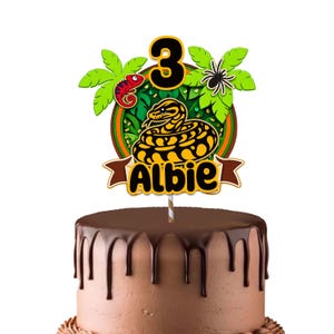 Peut inclure: Une décoration de gâteau sur le thème de la jungle avec un grand chiffre 3, un serpent jaune et noir enroulé, un caméléon rouge et une araignée. Le motif comprend des feuilles de palmier vertes et une bannière brune avec le nom Albie en noir.