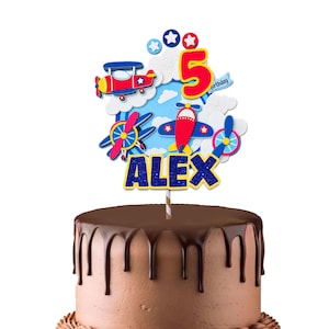 Pode incluir: Um topo de bolo com tema de aviões vermelhos e azuis, nuvens brancas, um número 5 grande em vermelho e o nome ALEX em letras azuis com glitter. O topo está montado num palito às riscas e colocado sobre um bolo com cobertura de chocolate.