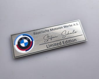 May include: A rectangular, brushed metal plaque featuring the BMW logo in a circular design with blue, white, red, and black colors. The text "Bayerische Motoren Werke A.G", "Stefan Schulz", and "Limited Edition" are also present.