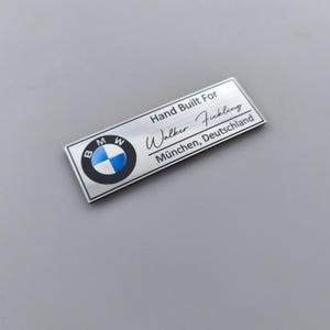 Könnte beinhalten: Ein rechteckiges, silberfarbenes Metallschild mit dem BMW-Logo auf der linken Seite. Der Text lautet Hand Built For Walker Fickling München, Deutschland. Die Oberfläche hat ein gebürstetes Metallfinish mit schwarzer Schrift und einem blau-weißen Logo.