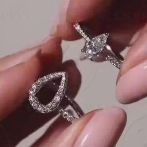 May include: A silver-toned ring featuring a pear-shaped setting encrusted with small clear stones. The band is thin and also adorned with small clear stones along the sides. The design includes a prominent teardrop-shaped focal point.