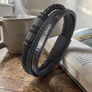 Pode incluir: Uma pulseira de couro preta com um design trançado e a inscrição "I love you always". A pulseira apresenta um fecho magnético e detalhes metálicos decorativos. É exibida em uma superfície de madeira ao lado de um livro e uma caneca.