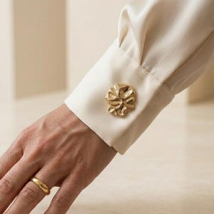 May include: A cream-colored long-sleeve shirt with a structured cuff featuring a large, irregular, gold-toned metallic button with a textured, organic surface. A simple, smooth gold-toned band ring is worn on the ring finger of the hand.