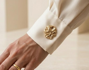 Puede incluir: Una camisa de manga larga color crema con un puño estructurado que presenta un botón metálico grande, irregular y de tono dorado con una superficie orgánica texturizada. Un anillo de banda dorado y liso se lleva en el dedo anular.