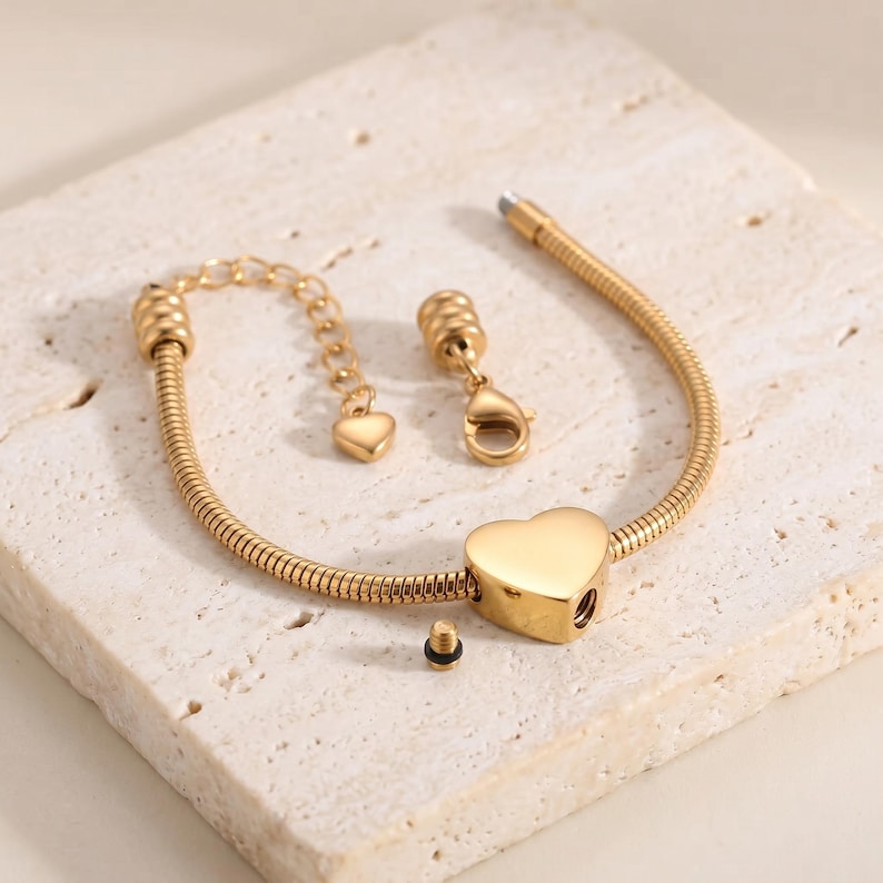 May include: A gold-toned snake chain bracelet featuring a central heart-shaped charm with a removable screw-top component. The bracelet includes an adjustable chain extension with a small heart-shaped end piece and a lobster claw clasp closure.