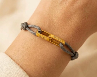 Puede incluir: Una pulsera de cordón gris con dos eslabones metálicos rectangulares dorados entrelazados. Cada eslabón está grabado con el texto S22.10.25 y M22.10.25 respectivamente. El cordón está asegurado con nudos a ambos lados de la pieza central.