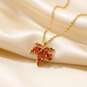Pode incluir: Um colar de tom dourado com um pingente em forma de folha de bordo. O pingente de folha é incrustado com várias pedras preciosas laranja-avermelhadas em corte marquise e redondo, fixadas numa armação de metal dourado e uma corrente fina.