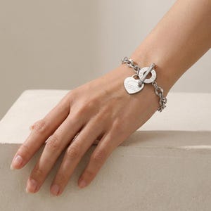 May include: A silver-toned chain link bracelet featuring a toggle clasp and a heart-shaped pendant. The circular toggle component is engraved with text, and the heart charm also displays small engraved lettering on its surface.