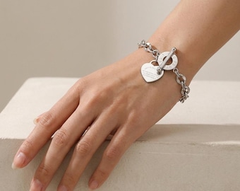 Puede incluir: Una pulsera de eslabones en tono plateado con un cierre de barra y un colgante en forma de corazón. El componente circular del cierre tiene texto grabado y el dije de corazón muestra un texto pequeño y legible en su superficie, creando un accesorio.