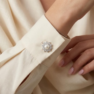 May include: A decorative button fastened to the cuff of a cream-colored long-sleeved shirt. The button features a central white pearl surrounded by a circular arrangement of small, clear, sparkling stones set in a silver-toned metal frame.