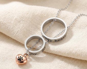 Puede incluir: Un collar de tono plateado con dos anillos entrelazados grabados con el texto "by my heart" y un pequeño dije de corazón en tono oro rosa que cuelga del anillo inferior, decorado con un gráfico de huella negra en su superficie.