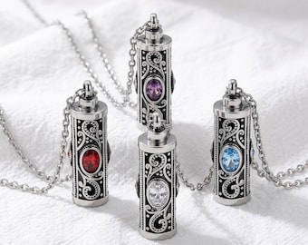 Puede incluir: Cuatro colgantes cilíndricos de metal en cadenas de tono plateado, cada uno con grabados intrincados de volutas negras y una piedra preciosa de corte ovalado en rojo, morado, transparente o azul claro, suspendidos de una pequeña argolla.