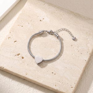 May include: A silver-toned snake chain bracelet featuring a smooth, polished heart-shaped charm at the center. The bracelet includes a lobster claw clasp and an adjustable extender chain finished with a tiny heart-shaped charm at the end.
