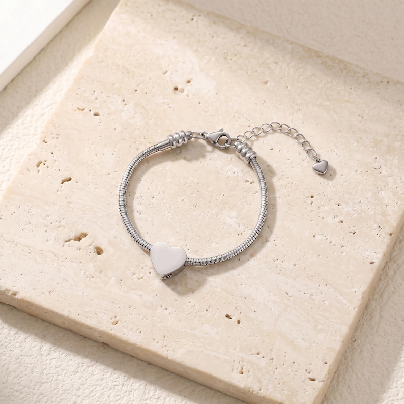 May include: A silver-toned snake chain bracelet featuring a smooth, polished heart-shaped charm at the center. The bracelet includes a lobster claw clasp and an adjustable extender chain finished with a tiny heart-shaped charm at the end.
