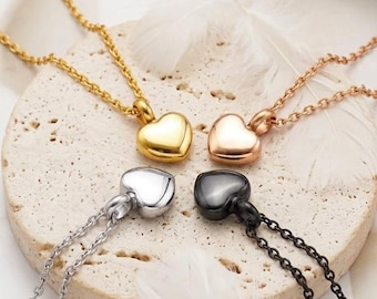 Puede incluir: Cuatro collares con colgantes en forma de corazón en oro, oro rosa, plata y negro. Cada colgante está suspendido de una delicada cadena. Los collares se exhiben sobre una superficie texturizada de color claro con plumas blancas.