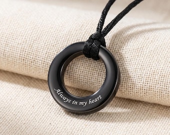 Puede incluir: Un colgante circular negro suspendido de un cordón negro. El colgante presenta un acabado pulido y está grabado con el texto cursivo blanco Always in my heart. El diseño es sencillo y cuenta con una estructura de anillo de metal liso.