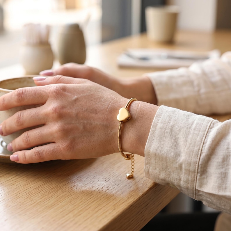 May include: A gold-toned snake chain bracelet featuring a smooth, solid heart-shaped charm. The bracelet includes an adjustable extender chain that terminates in a small, dangling heart-shaped charm, providing a secure and flexible fit.