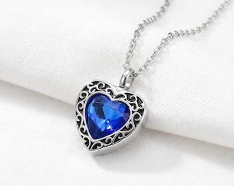 Puede incluir: Un collar de tono plateado con un colgante en forma de corazón que presenta una piedra preciosa facetada de color azul intenso en el centro. El colgante está enmarcado por intrincados patrones de filigrana metálica y cuelga de una cadena.