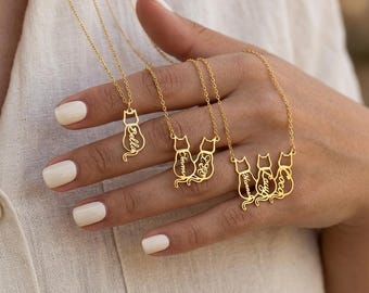 Peut inclure: Trois colliers dorés avec des pendentifs en forme de chat. Un pendentif porte le nom Bella, un autre les noms Emma et Leo, et le troisième présente les noms Noma, Lucy et Coco intégrés dans les silhouettes des chats.