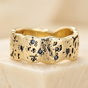 May include: A wide gold band ring with a textured, cratered surface finish. The ring features several small, round, deep blue gemstones embedded within the irregular, pitted indentations across the outer surface of the metal band.