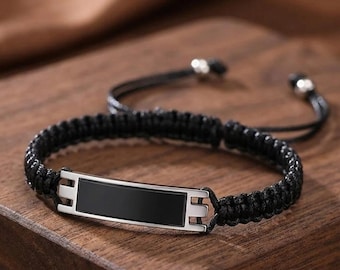 Puede incluir: Una pulsera de cordón trenzado negro con una placa central rectangular de metal plateado con una incrustación negra lisa. La pulsera incluye cordones ajustables terminados con pequeñas cuentas de metal plateado en los extremos de los cordones.