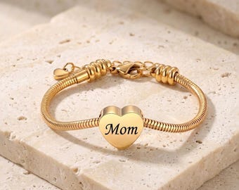 May include: A gold-toned snake chain bracelet featuring a central heart-shaped charm engraved with the word Mom. The bracelet includes a lobster claw clasp, decorative ribbed metal segments near the closure, and a small dangling heart charm.