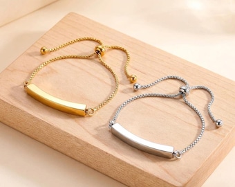 Op de afbeelding: Twee verstelbare armbanden met een gladde, gebogen metalen staaf als middelpunt. De ene armband heeft een gepolijste goudkleurige afwerking, terwijl de andere een gepolijste zilverkleurige afwerking heeft. Beide gebruiken een fijne boxketting.