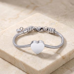 May include: A silver-toned snake chain bracelet featuring a central heart-shaped charm. The bracelet includes a lobster claw clasp, an adjustable extender chain, and a small dangling heart charm at the end of the extension for a secure fit.