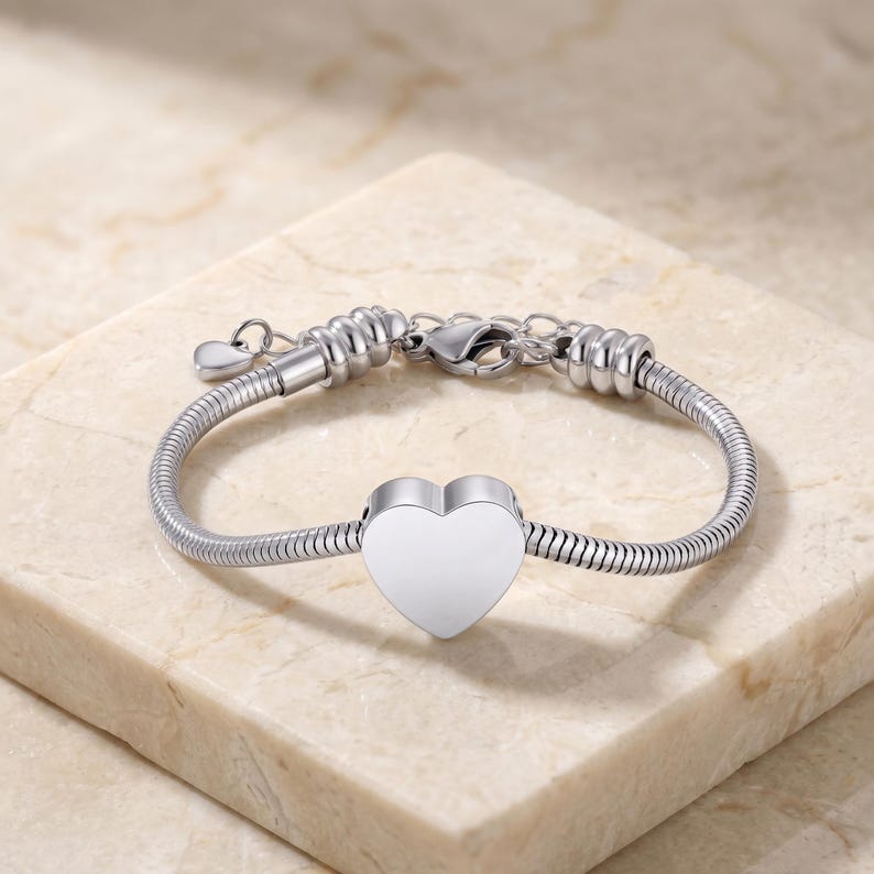 May include: A silver-toned snake chain bracelet featuring a central heart-shaped charm. The bracelet includes a lobster claw clasp, an adjustable extender chain, and a small dangling heart charm at the end of the extension for a secure fit.