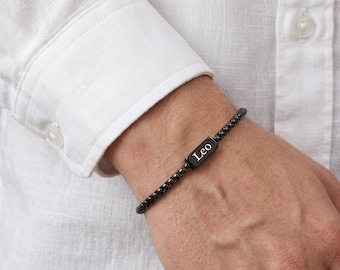 Puede incluir: Una pulsera de cadena tipo caja negra con un dije metálico rectangular que tiene el nombre Leo grabado en letras blancas. La pulsera está construida con pequeños eslabones cuadrados uniformes que forman una banda flexible y continua.
