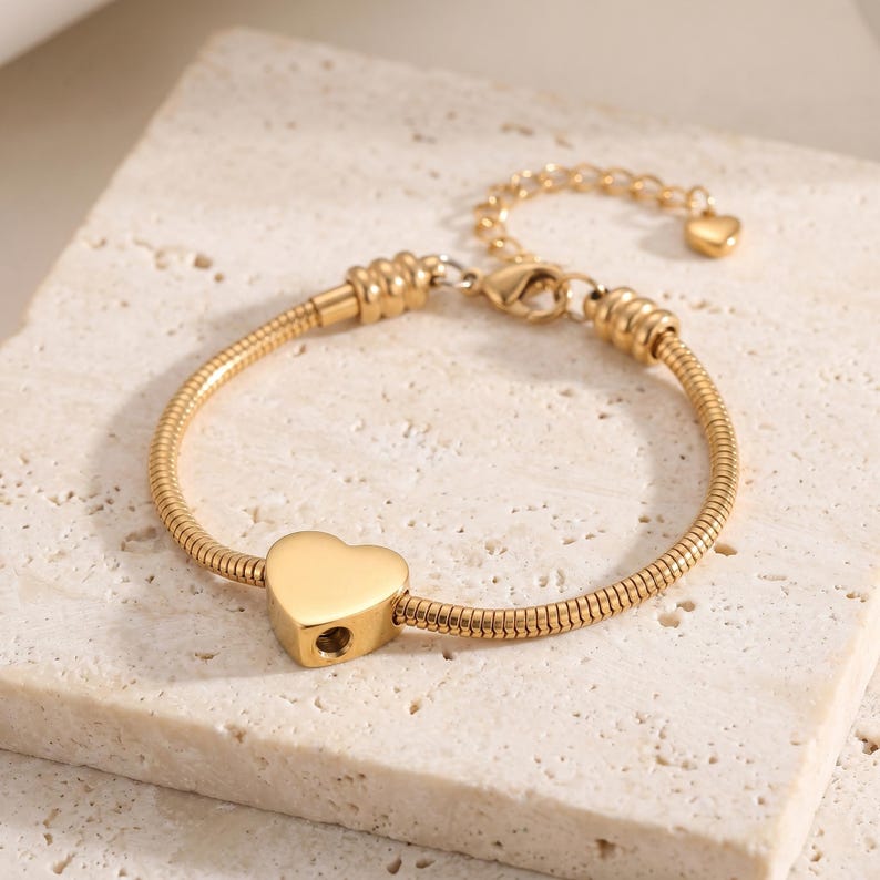 May include: A gold-toned snake chain bracelet featuring a central heart-shaped charm with a small circular opening on the side. The bracelet includes a lobster claw clasp, a short extender chain, and a tiny heart-shaped charm at the end of the chain.