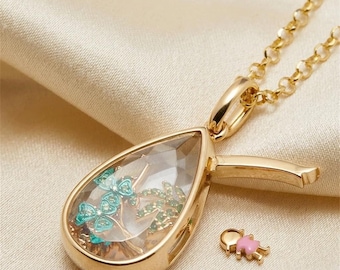 Puede incluir: Un colgante de relicario en forma de lágrima de tono dorado con una cadena a juego. El relicario tiene un panel frontal transparente que revela dijes internos en forma de árbol verde y dorado. Una pequeña figura dorada y rosa descansa al lado.
