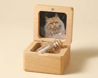 Puede incluir: Una caja de madera de color claro con tapa abatible que presenta una fotografía cuadrada de un gato naranja esponjoso. Dentro de la base abierta de la caja hay un pequeño frasco de vidrio transparente con un tapón de corcho.