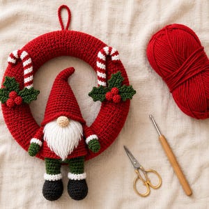 May include: A red crocheted wreath featuring a gnome with a white beard, red hat, green sleeves, and black boots. The wreath is decorated with two candy canes and holly leaves with red berries. A ball of red yarn, a crochet hook, and scissors.