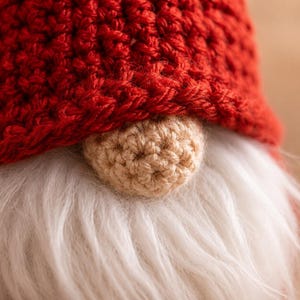 May include: A close-up of a gnome figurine featuring a textured, deep red crocheted hat that covers the upper portion of the face. A small, round, beige crocheted nose sits prominently above a thick, soft, white faux fur beard.