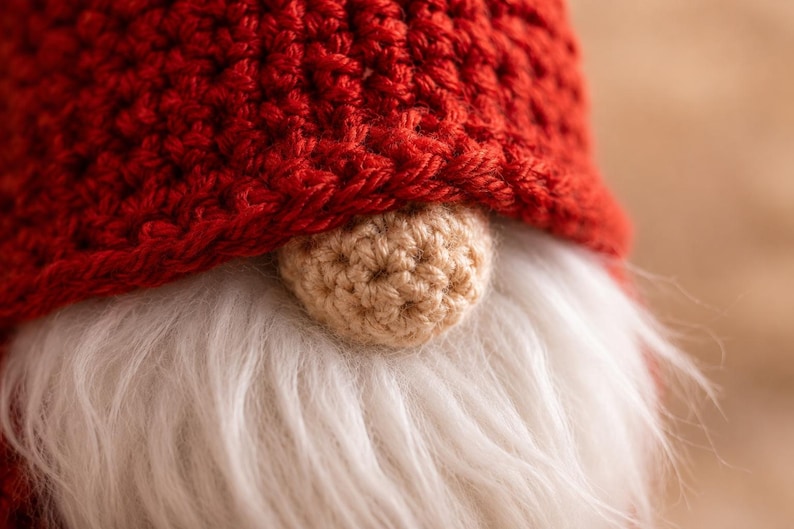May include: A close-up of a gnome figurine featuring a textured, deep red crocheted hat that covers the upper portion of the face. A small, round, beige crocheted nose sits prominently above a thick, soft, white faux fur beard.