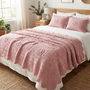 May include: A dusty rose crochet bedspread and two matching square throw pillows featuring intricate floral patterns and scalloped edges. The textile set is draped over a bed with white linens, showcasing a detailed, textured knit construction.