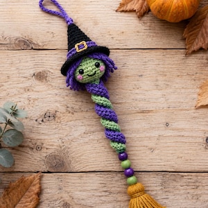 Può includere: Un ornamento a forma di strega all'uncinetto con testa verde, occhi neri e guance rosa, che indossa un cappello nero con fibbia gialla. Il corpo è una spirale viola e verde, rifinita con perline e una nappa in filato giallo senape.