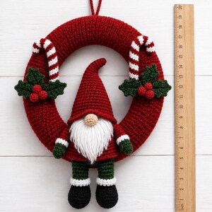 May include: A red crocheted wreath featuring a central gnome with a white beard, red hat, and green trousers. The wreath is decorated with two candy canes and holly leaves with red berries. A wooden ruler shows the wreath is 38 cm in diameter.