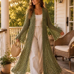 Può includere: Un cardigan lungo fino ai piedi in crochet verde oliva, aperto sul davanti, con maniche lunghe svasate e bordi smerlati, indossato sopra una canotta bianca a coste e pantaloni ampi color crema, accompagnato da una borsa rotonda in paglia.