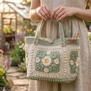 Puede incluir: Un bolso de ganchillo en color verde salvia y crema con un motivo cuadrado que presenta una margarita blanca central y hojas verdes. El bolso tiene dos asas resistentes, un cierre de botón de madera y una etiqueta de cuero con texto.
