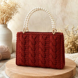 May include: A deep red rectangular handbag featuring a prominent cable knit texture across the front. The bag is equipped with a single arched handle composed of smooth, round white faux pearls attached to the body with gold-toned metal rings.