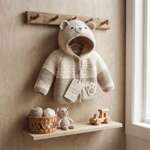 May include: A beige and cream crochet hooded jacket featuring bear ears on the hood, two wooden buttons, and a paw print applique. A white tag labeled Handmade by Pitter Patter hangs from the front. A small crochet bear and wooden toy train sit below.