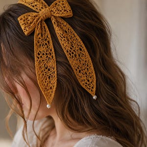 May include: A mustard yellow crochet headband featuring an intricate lace pattern and a large bow detail. Two long, pointed fabric tails extend from the bow, each finished with a small, round white pearl bead at the tip, creating a textured accessory.