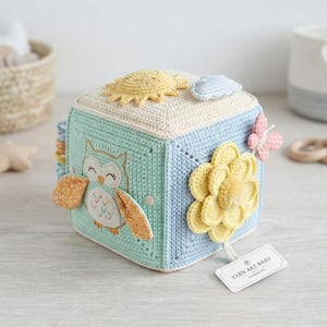 May include: A crocheted sensory cube featuring a mint green side with an embroidered owl, a light blue side with a large yellow flower and a pink butterfly, and a cream top with a yellow sun and blue cloud. A white tag reads YARN ART BABY.