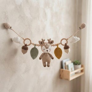 May include: A crochet baby stroller toy featuring a central deer figure, two wooden rings, two acorns, two leaves in olive and mustard, and two white clouds, all strung on a cord with small wooden beads and secured by two metal clips at the ends.
