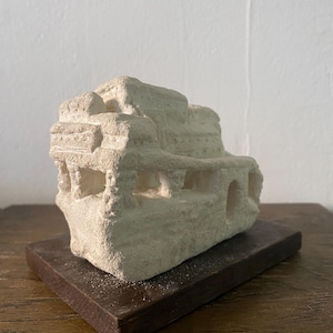 May include: A beige, carved stone sculpture of a building with architectural details, set on a dark brown rectangular base. The sculpture has multiple levels and openings, resembling a miniature ancient structure.