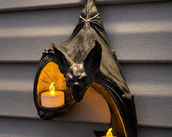 May include: A dark gray wall-mounted candle holder shaped like a hanging bat with textured wings. The bat features two circular platforms designed to hold tea light candles, each containing a small, glowing, battery-operated white tea light.