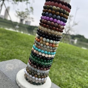 Op de afbeelding: Een display van kralenarmbanden in diverse kleuren, waaronder rood, bruin, paars, zwart, turquoise en groen. De armbanden zijn gestapeld op een grijze cilindrische standaard met een witte basis, met een verscheidenheid aan edelsteen- en kralenontwerpen.